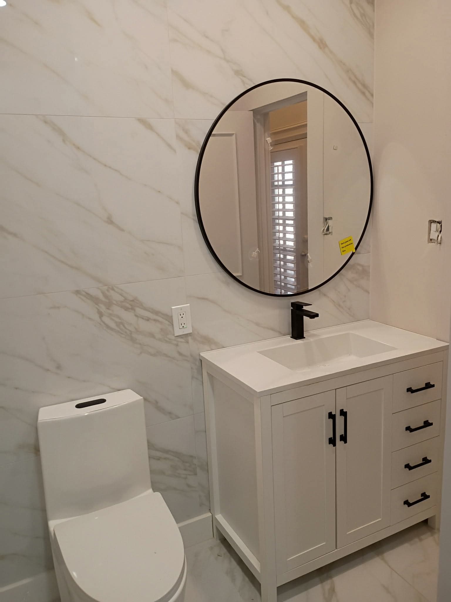 Modern bathroom with white marble walls, a round black mirror, and a white vanity.