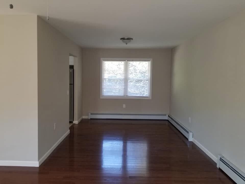 Empty room with dark hardwood floors, beige walls, a window, and white baseboard heaters.