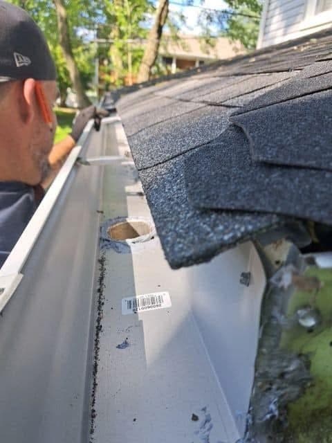 Man in a baseball cap inspects a newly installed white gutter on a shingled roof.