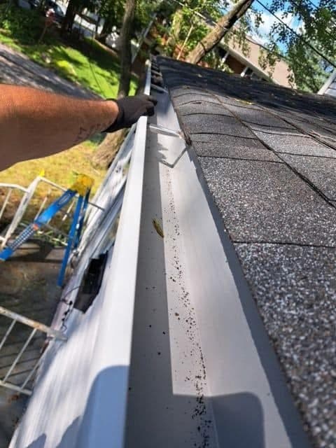 A gloved hand inspects a clean white gutter attached to a dark shingled roof.