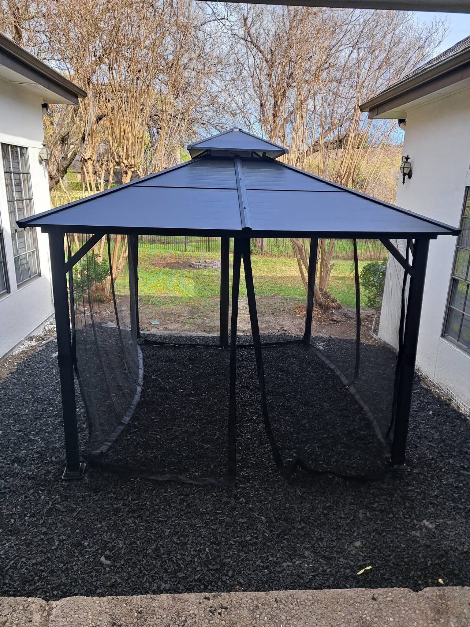 Black metal gazebo with tiered roof and mesh netting on dark mulch between two buildings.
