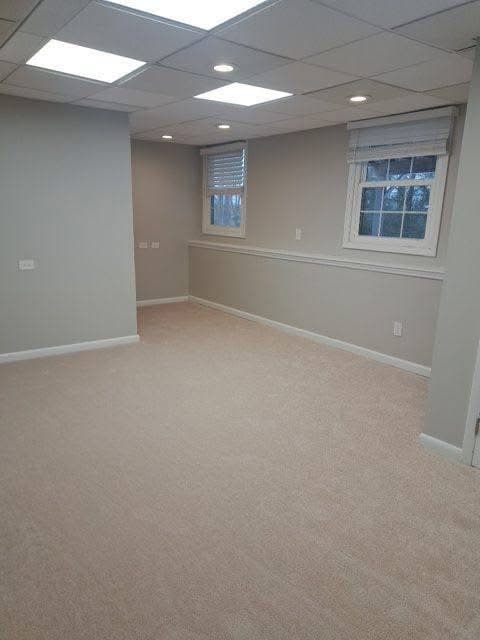 Empty finished basement with beige carpet, light gray walls, and bright overhead panel lighting.