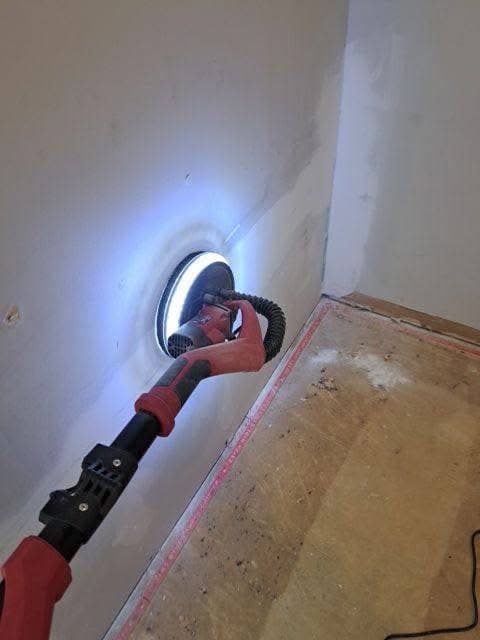 Red drywall sander with a circular LED light ring smoothing an unfinished wall surface.